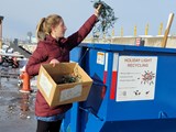 A person holding a cardboard box with holiday lights taking them out and placing them in a blue dumpster labeled holiday light recycling