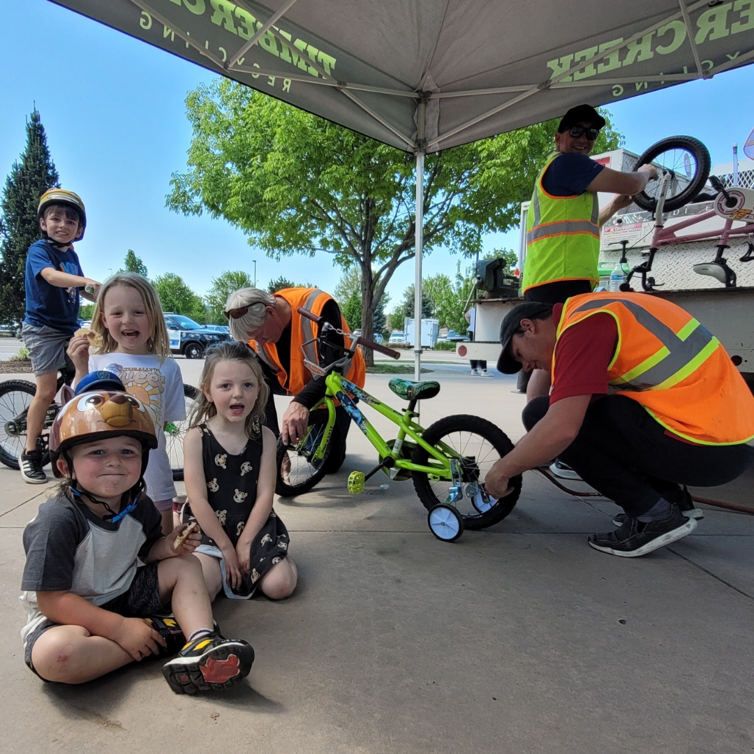 Volunteers in safety vests working on a green kid's bike while four kids pose for the camera